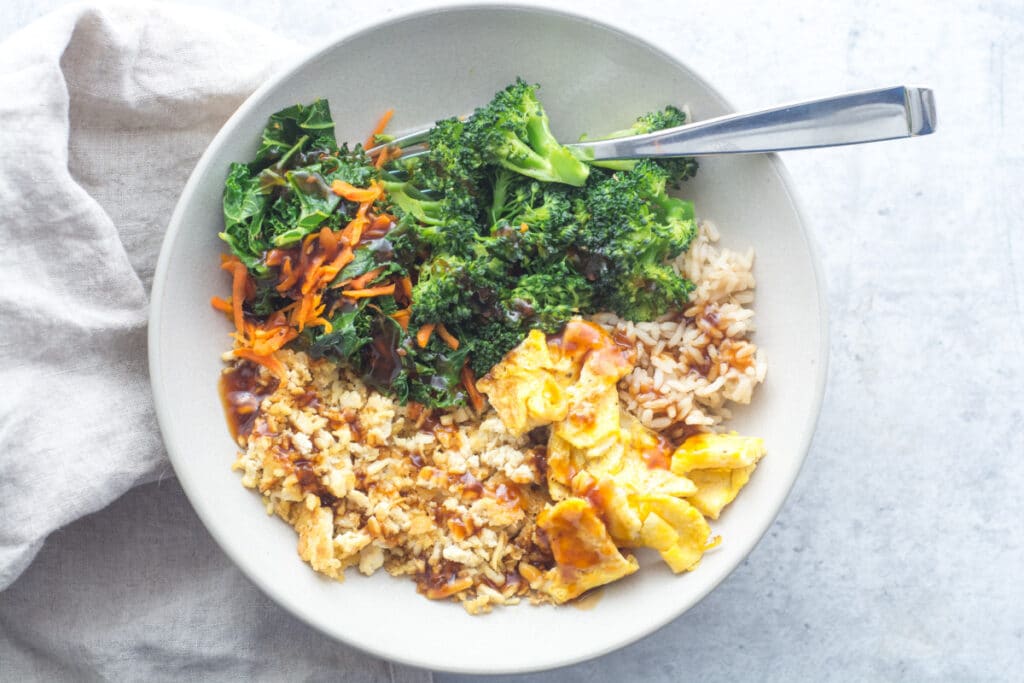 A bowl with tofu, rice, vegetables, and eggs sits on a gray surface with a fork and a napkin.